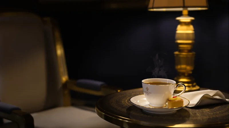 Steaming teacup on saucer at a dark table, empty chair in background, gold lamp and folded napkin in candlelight