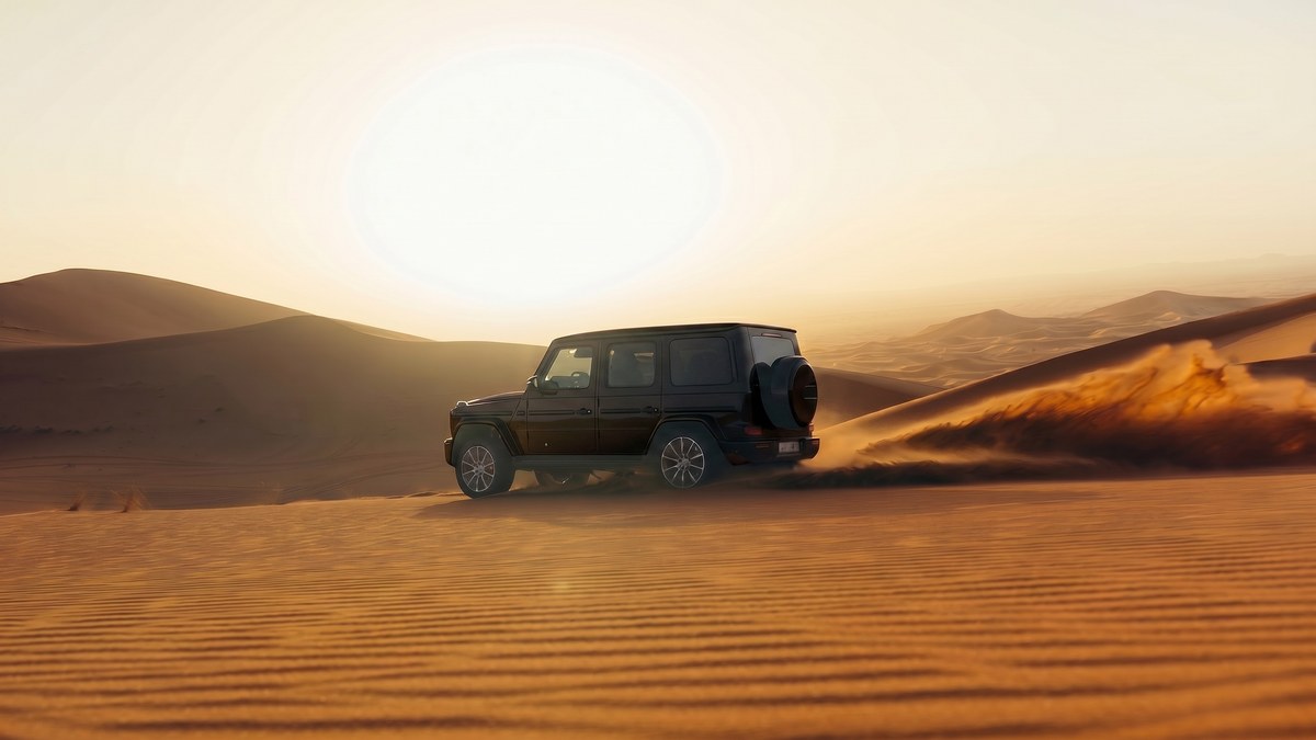A black luxury SUV in distant three quarter rear profile cresting a desert dune at golden hour with sand kicking up from its rear tyres